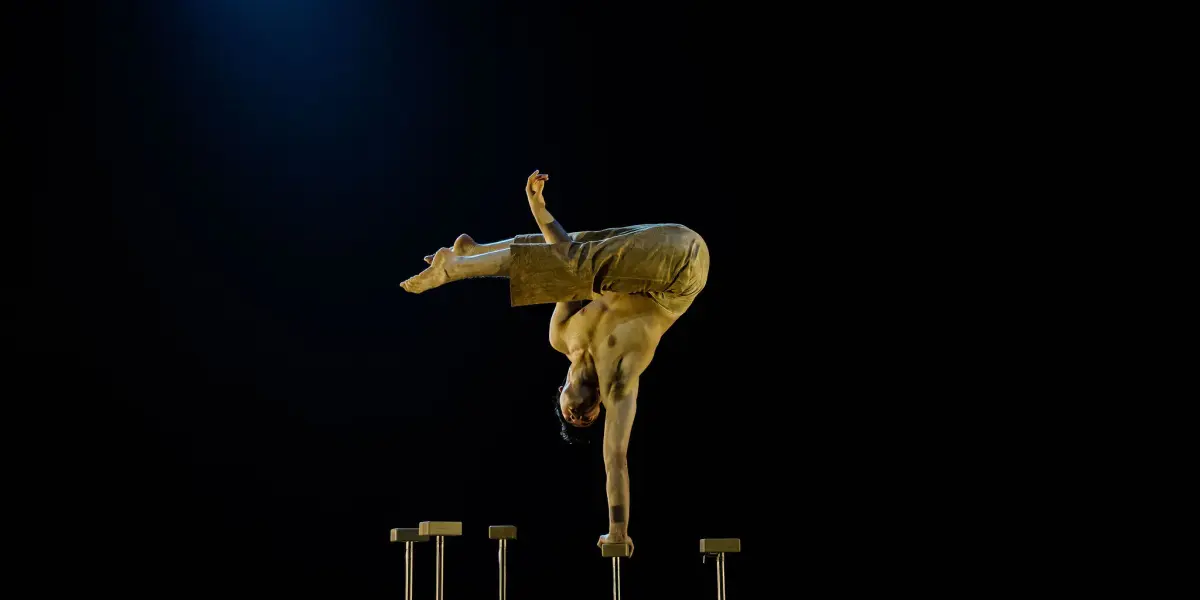Maël Trân se tient en équilibre sur une main au sommet d'un grand support, le corps plié et les jambes tendues en l'air, réalisant un remarquable équilibre sur une toile de fond sombre à l'éclairage dramatique.