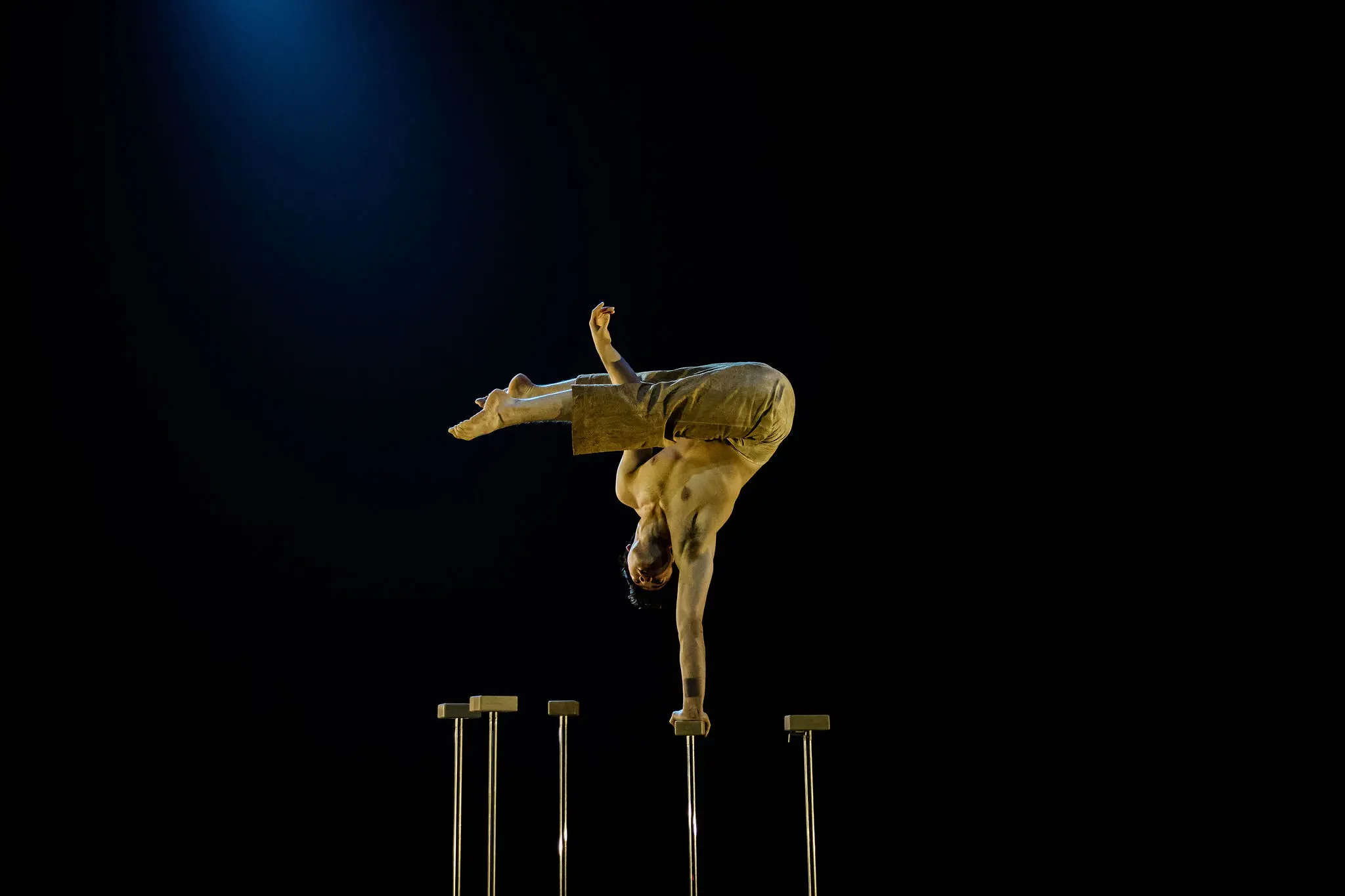 Maël Trân se tient en équilibre sur une main au sommet d'un grand support, le corps plié et les jambes tendues en l'air, réalisant un remarquable équilibre sur une toile de fond sombre à l'éclairage dramatique.