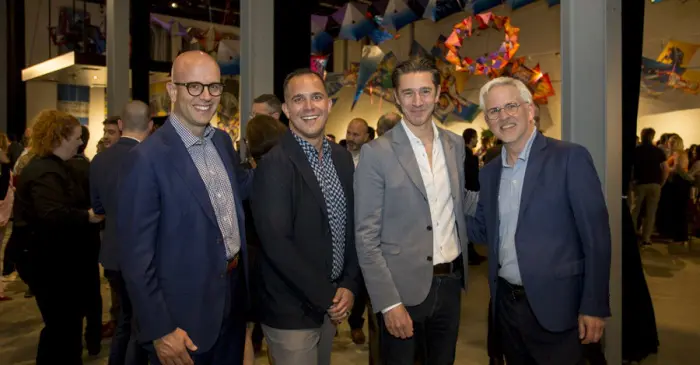 Quatre hommes en costume sourient et posent ensemble lors d'un événement en salle, célébrant un don d'un million de dollars du Groupe Cirque du Soleil, avec des décorations colorées ressemblant à des cerfs-volants et d'autres participants en arrière-plan.
