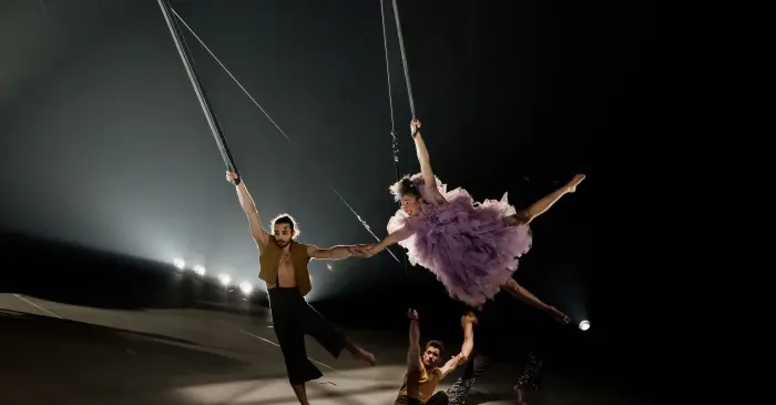 Deux artistes aériens en costume, dont l'un porte une robe violette, sont suspendus à des trapèzes et se tiennent par la main au-dessus d'une scène faiblement éclairée - une scène inspirante rendue possible grâce à l'implication et au soutien du Groupe FlightHub.