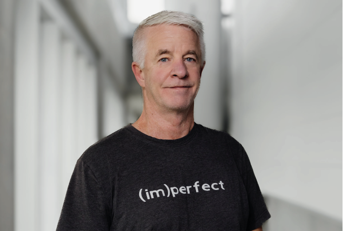 Un homme d'âge moyen aux cheveux gris courts se tient à l'intérieur, face à la caméra. Il porte un T-shirt sombre sur lequel on peut lire (im)perfect en texte blanc. L'arrière-plan légèrement flou révèle un couloir moderne, évoquant le style minimaliste de Marianella Michaud.