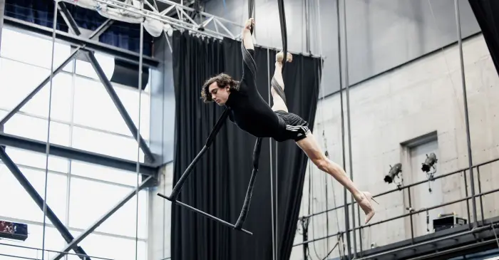 Une personne effectue un numéro aérien en intérieur, suspendue horizontalement à un tissu noir et à une barre de trapèze, un bras tendu et les jambes allongées, dans un studio spacieux, alors que l'ENC ouvre ses portes lors des Journées de la culture.