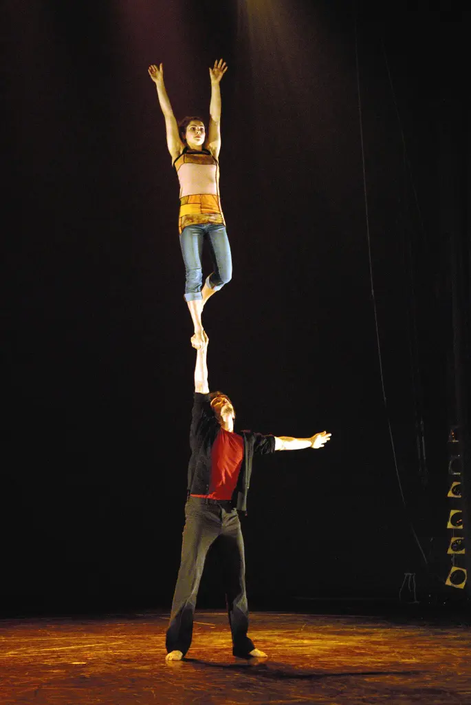 Sur une scène faiblement éclairée, Justine Méthé-Crozat se tient en équilibre sur un pied au dessus de la main tendue de Philippe Renaud, les deux bras levés, faisant preuve d'une force et d'une coordination remarquables.