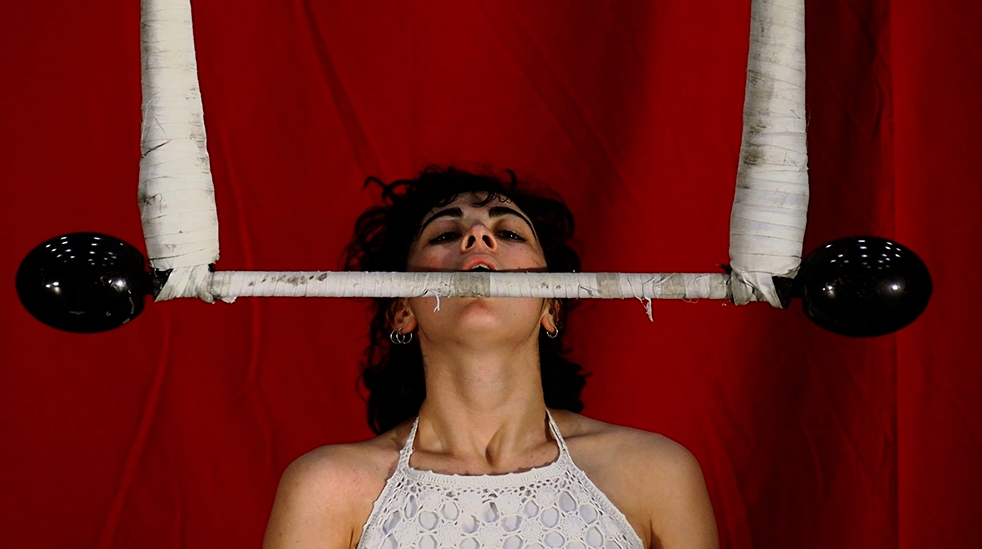 Au festival CIRCA d'Auch, une personne aux cheveux noirs bouclés tient une barre de trapèze en équilibre sous le menton, sur un fond rouge vif. Elle porte un dos nu blanc en crochet et affiche une expression concentrée.