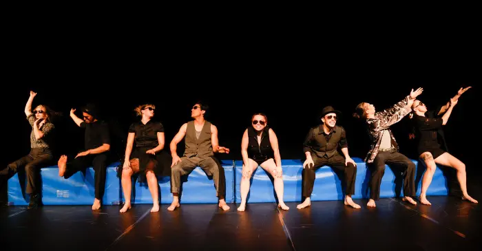 Huit personnes pieds nus, vêtues de costumes sombres et portant des lunettes de soleil, sont assises sur un banc bleu sur la scène, prenant des poses expressives et théâtrales sur un fond noir, dans l'esprit avant-gardiste du festival du cirque actuel d'Auch.