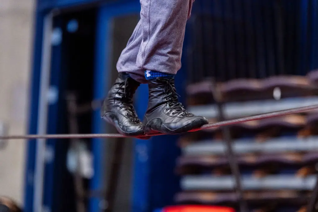 Une personne portant des bottes noires et un pantalon bleu marche prudemment sur une corde raide, avec des sièges flous et un fond bleu visible derrière elle, incarnant l'équilibre et la vigilance d'un protecteur national de l'élève.
