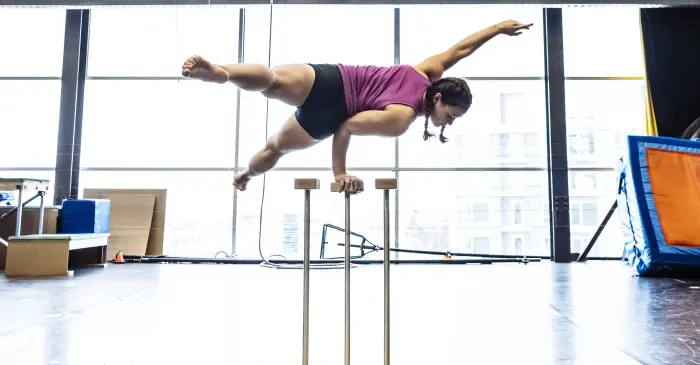 Dans un studio lumineux et spacieux, une personne en tenue de sport se tient en équilibre sur une main au dessus de grandes cannes d'équilibre, mettant en valeur les compétences que le Concours Élève d'un jour permet à l'ENC d'accueillir des 9 à 17 ans a pour but d'inspirer.