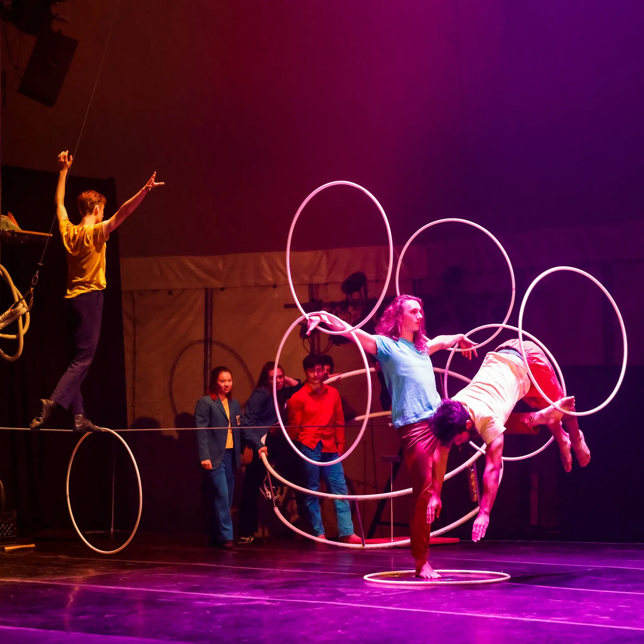Cinq artistes de cirque entrent en scène dans un préambule vibrant ; l'un se tient en équilibre sur une corde raide tandis que les autres interagissent avec de grands cerceaux, leurs costumes colorés et leurs éclairages dramatiques créant une atmosphère dynamique et animée.