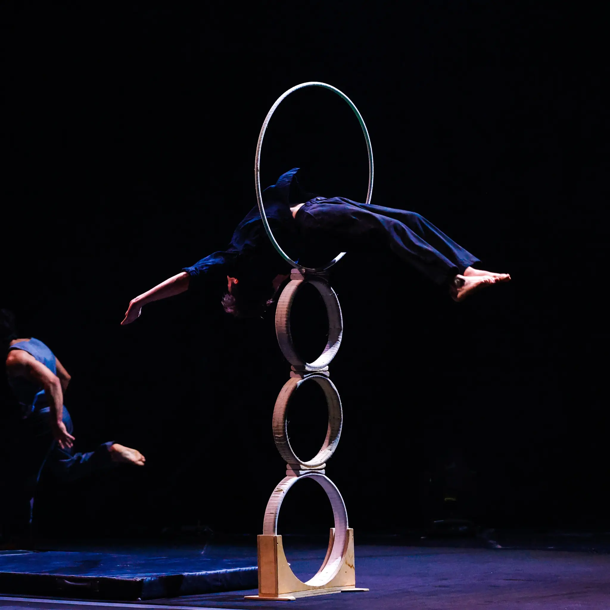 Un artiste vêtu de vêtements sombres se penche en arrière dans un grand cerceau posé sur trois anneaux empilés sur scène, évoquant l'art de Triptyque, tandis qu'une autre personne se déplace à l'arrière-plan sur une toile de fond sombre.