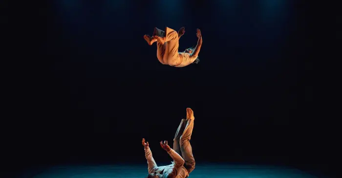 Deux acrobates évoluent sur une scène sombre sous les projecteurs, leurs costumes beiges reflétant la subvention accordée au projet N.Ormes pour favoriser l'inclusion et la diversité au sein des arts vivants, tandis que l'un se tient en équilibre dans les airs au-dessus de l'autre.