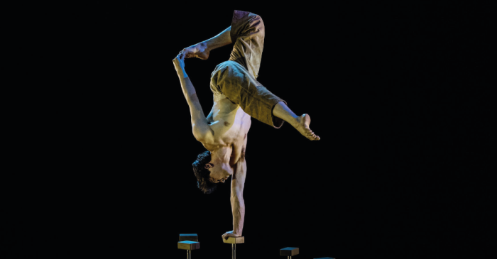 Un acrobate torse nu se tient en équilibre sur une main au sommet d'un support, les jambes pliées et un pied touchant sa tête, exécutant une pose de contorsion avancée des arts du cirque sur un fond sombre.