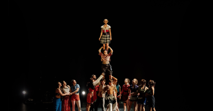 Un groupe d'artistes forme une pyramide humaine sur une scène sombre à Kaboum, avec une jeune fille en robe à carreaux en équilibre au sommet tandis que d'autres la soutiennent et la regardent d'en bas. Des projecteurs lumineux éclairent cette scène captivante.