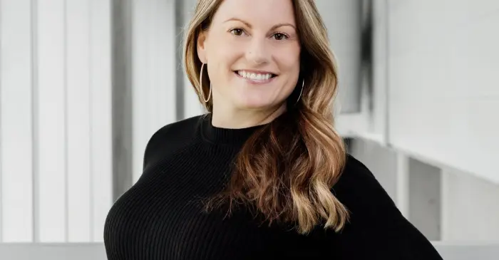 Une femme aux longs cheveux blonds ondulés, vêtue d'un pull noir, se tient à l'intérieur près d'une balustrade, souriant à la caméra dans un espace moderne et lumineux, célébrant la nomination de Marilou Cousineau au poste de directrice pédagogique.