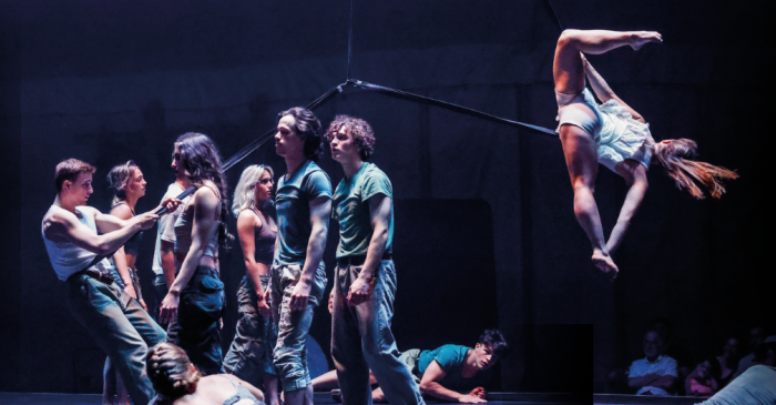 Un groupe d'acrobates de Kaboum se tient serré sur la scène, tandis qu'un artiste en blanc est suspendu à un poteau au-dessus d'eux, la tête en bas. La scène, très éclairée, contraste avec l'arrière-plan sombre.