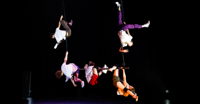 Cinq artistes de kaboum sont suspendus à des cordes ou à des équipements aériens sur un fond noir, certains à l'envers, d'autres s'agrippant avec les mains, tous vêtus de vêtements décontractés.