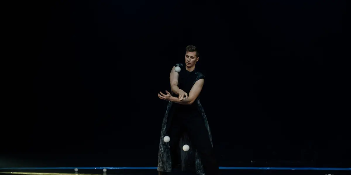 Pierre Domis, un artiste pieds nus vêtu d'une tenue sombre sans manches, jongle avec de multiples balles blanches sur une scène faiblement éclairée, certaines balles étant suspendues dans les airs et d'autres éparpillées sur le sol.