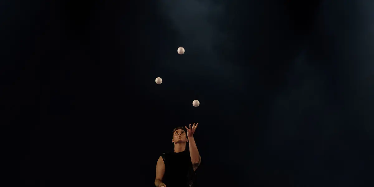 Pierre Domis, vêtu de noir, jongle avec de multiples balles blanches sur une scène sombre, éclairée par deux projecteurs en hauteur. Quelques balles sont éparpillées sur le sol autour de lui.