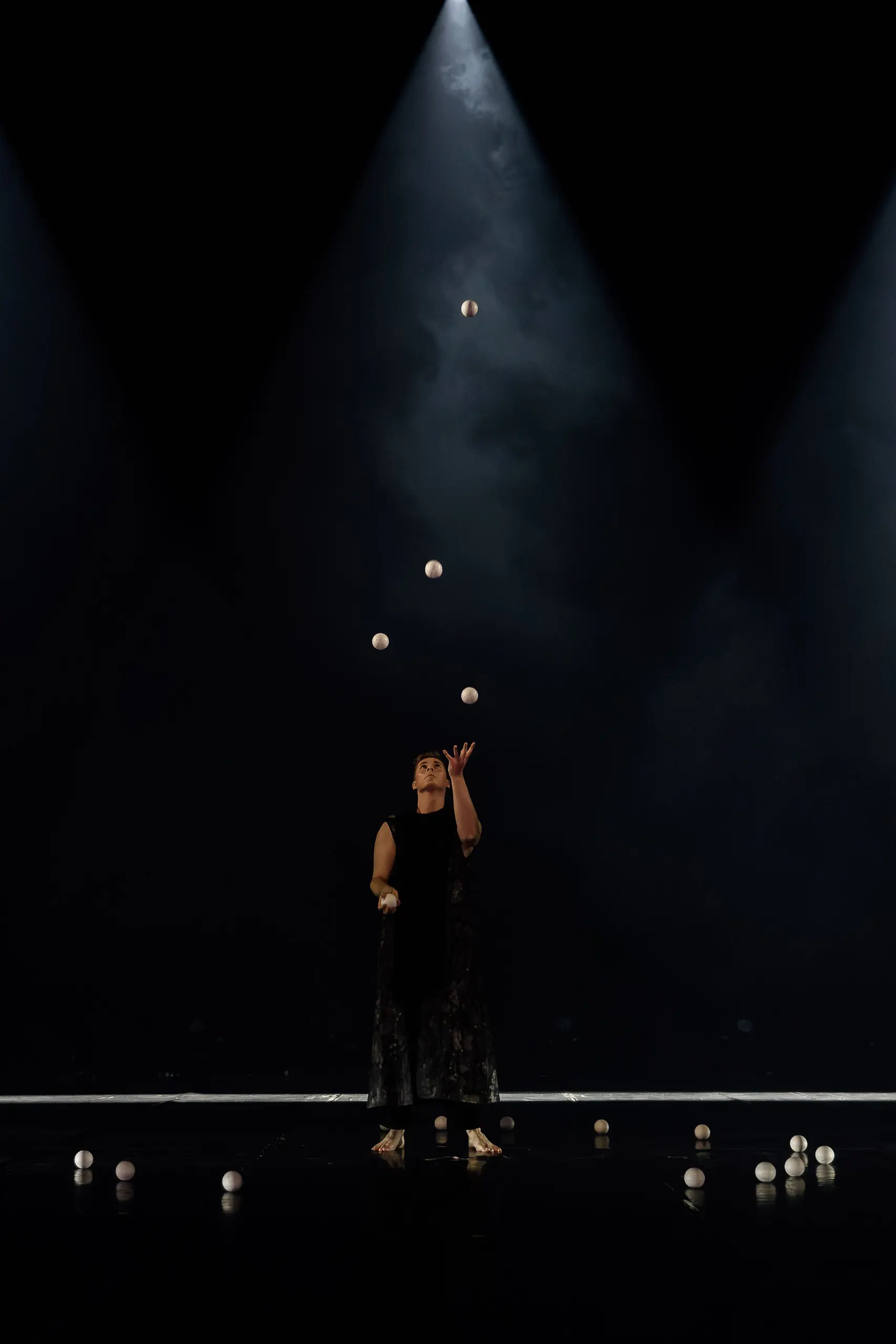 Pierre Domis, vêtu de noir, jongle avec de multiples balles blanches sur une scène sombre, éclairée par deux projecteurs en hauteur. Quelques balles sont éparpillées sur le sol autour de lui.