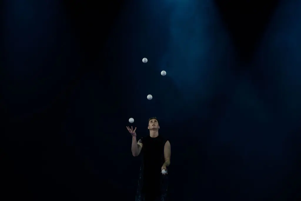 Pierre Domis, vêtu d'une tenue noire sans manches, jongle avec cinq balles blanches sous un éclairage bleu dramatique, seul sur une scène sombre.