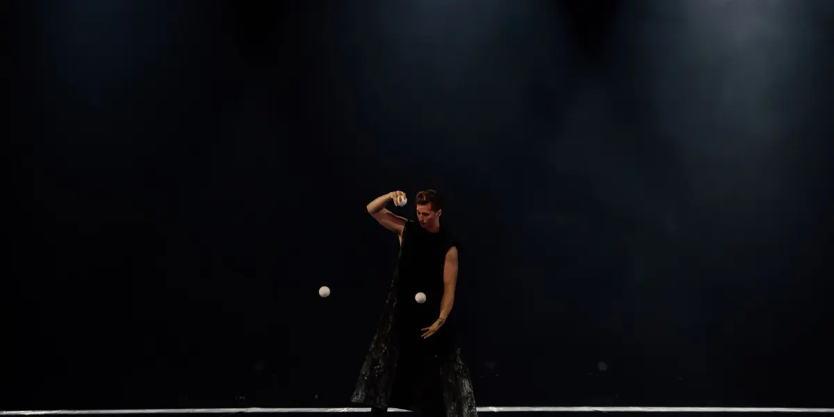 Une personne vêtue de vêtements sombres, rappelant Pierre Domis, jongle avec plusieurs balles blanches sur une scène faiblement éclairée, plusieurs balles étant éparpillées sur le sol autour d'elle.