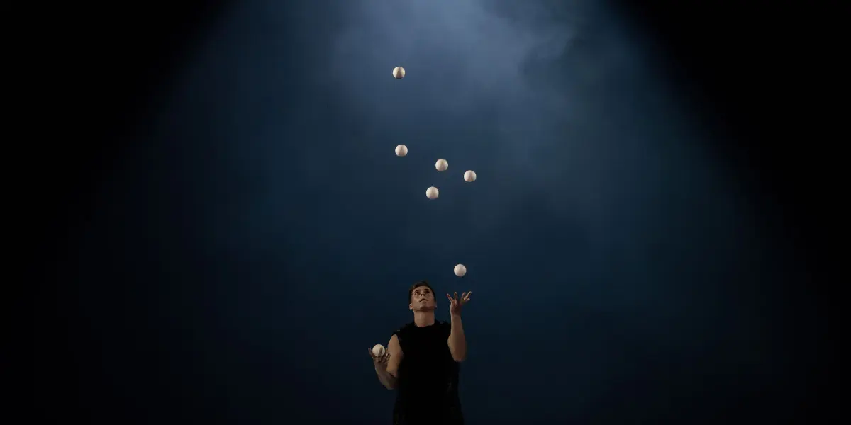 Pierre Domis, vêtu de noir, jongle avec sept balles blanches sous un projecteur dramatique, sur un fond sombre et enfumé.