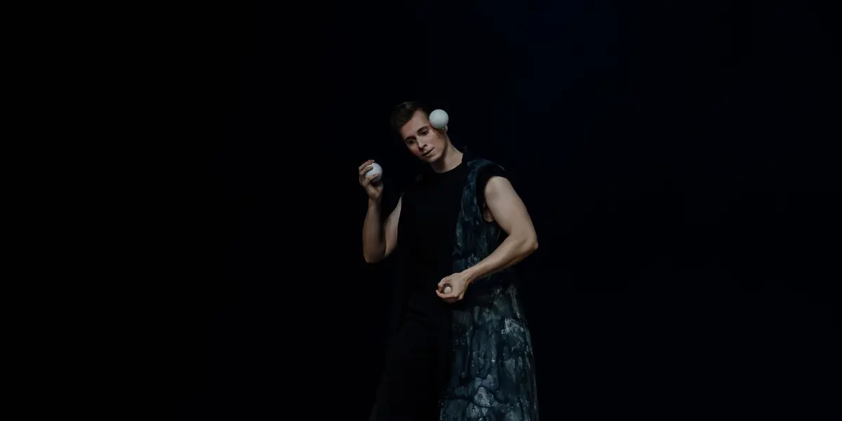 Pierre Domis, vêtu de noir et d'un long gilet à motifs, jongle avec des balles blanches sur une scène sombre, sous un éclairage dramatique.