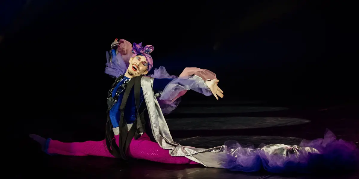 Aubin Constanceau, dans un costume coloré et fantaisiste, fait un grand écart sur scène, les bras levés et la bouche ouverte en signe d'expression. Il porte des collants rose vif, une longue cape fluide et un maquillage de scène spectaculaire.