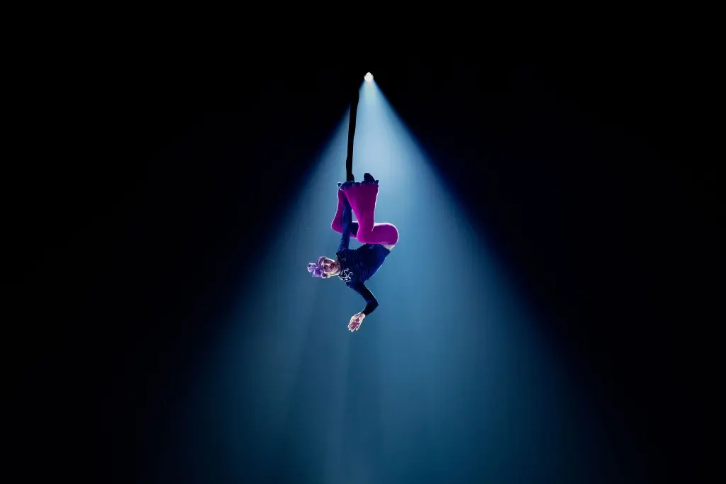 Aubin Constanceau, un artiste aérien vêtu d'un pantalon rose vif, est suspendu à une corde, la tête en bas, éclairé par un projecteur sur un fond sombre.