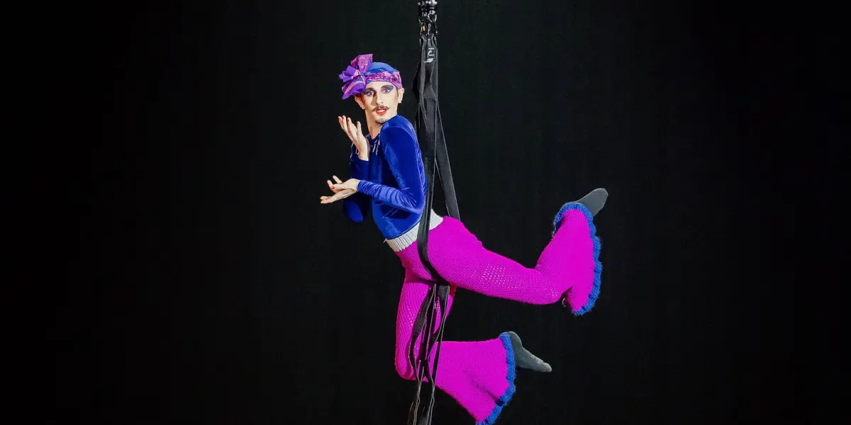 Aubin Constanceau, un artiste aérien vêtu d'un costume bleu et magenta vibrant, est suspendu à des soies noires, prenant une pose dramatique sous un projecteur sur un fond sombre.