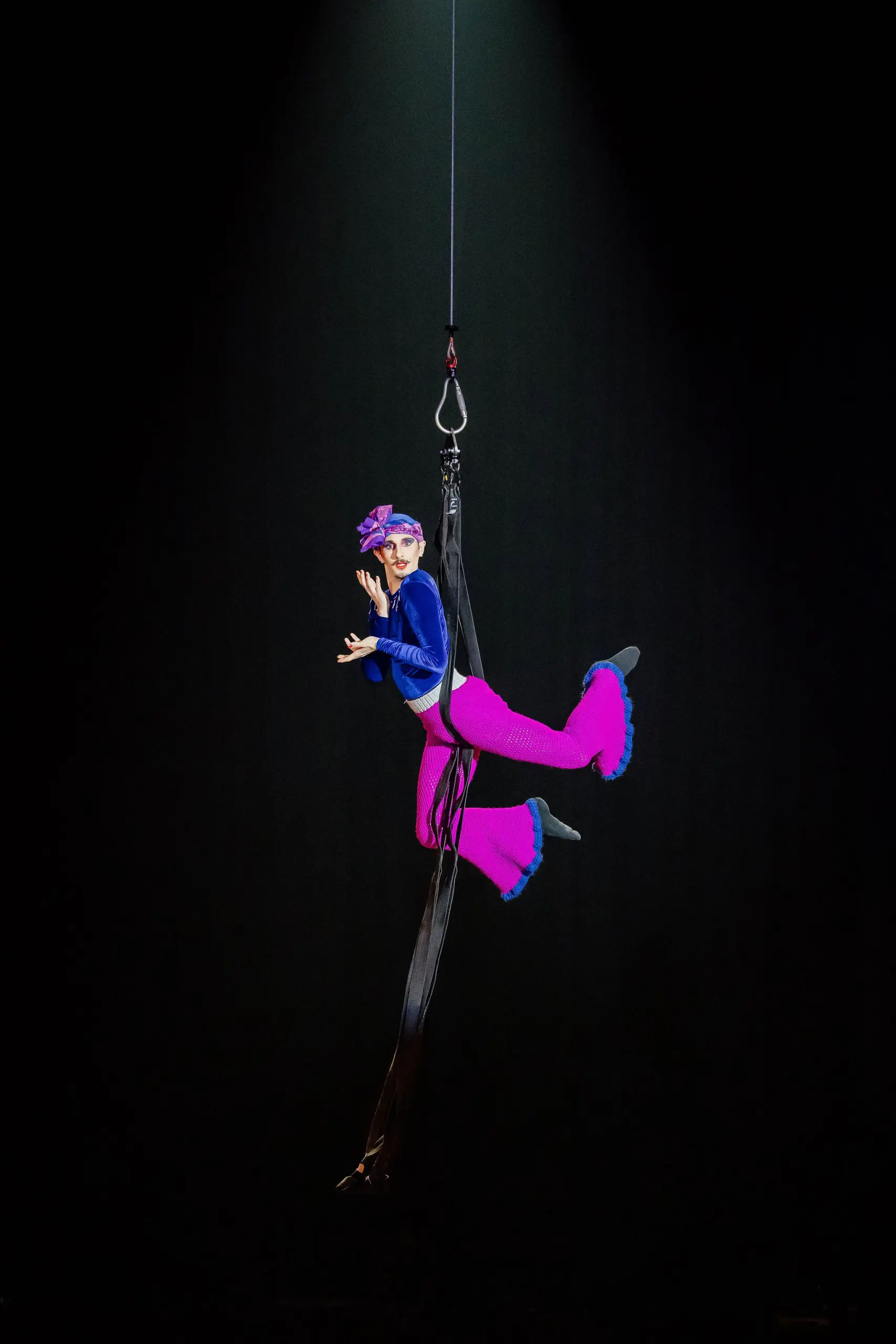 Aubin Constanceau, un artiste aérien vêtu d'un costume bleu et magenta vibrant, est suspendu à des soies noires, prenant une pose dramatique sous un projecteur sur un fond sombre.