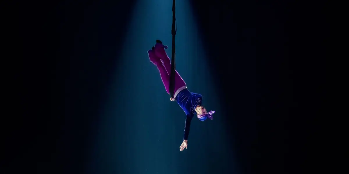 Aubin Constanceau, artiste aérien en tenue violette et foulard, est suspendu à l'envers sur une corde, éclairé par les projecteurs sur une scène sombre, avec un bras tendu vers le bas.