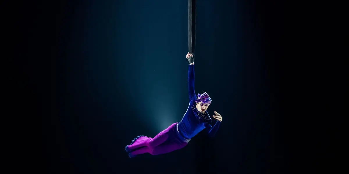 Aubin Constanceau, vêtu d'un pantalon violet vif et d'un haut bleu, est suspendu par un bras à une corde verticale, sous la lumière d'un projecteur sur une scène sombre.