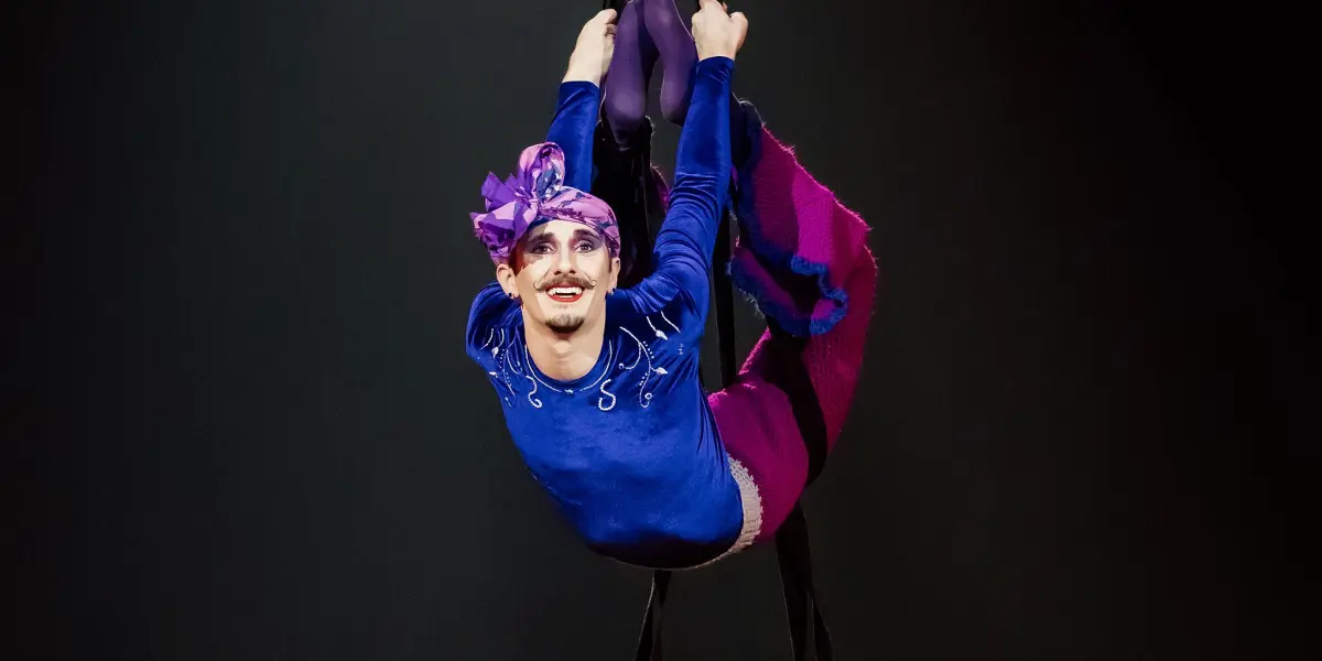 Aubin Constanceau, vêtu d'un vêtement coloré et d'une coiffe violette, est suspendu à des sangles, souriant, les jambes repliées, sous la lumière d'un projecteur, sur un fond sombre.