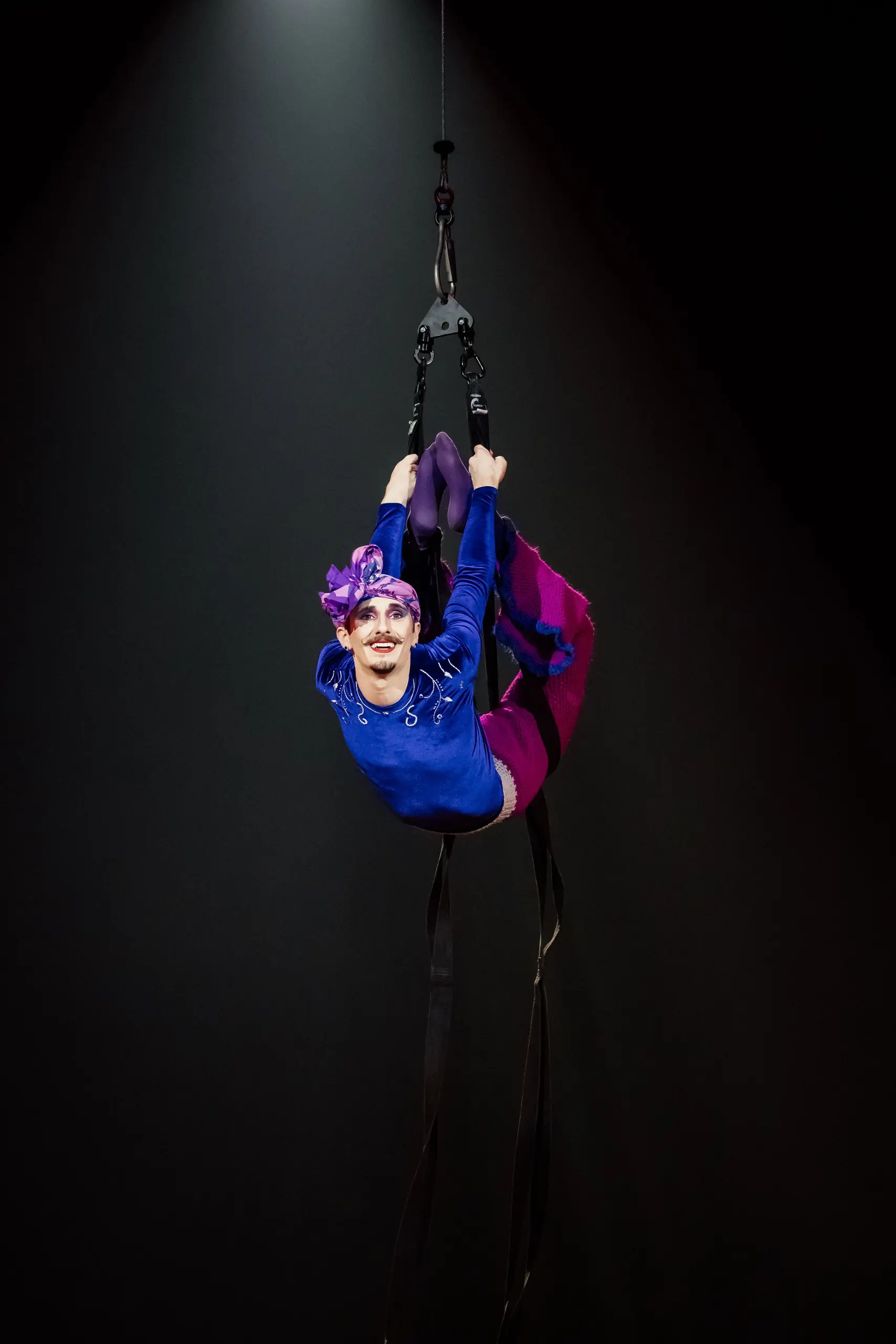 Aubin Constanceau, vêtu d'un vêtement coloré et d'une coiffe violette, est suspendu à des sangles, souriant, les jambes repliées, sous la lumière d'un projecteur, sur un fond sombre.