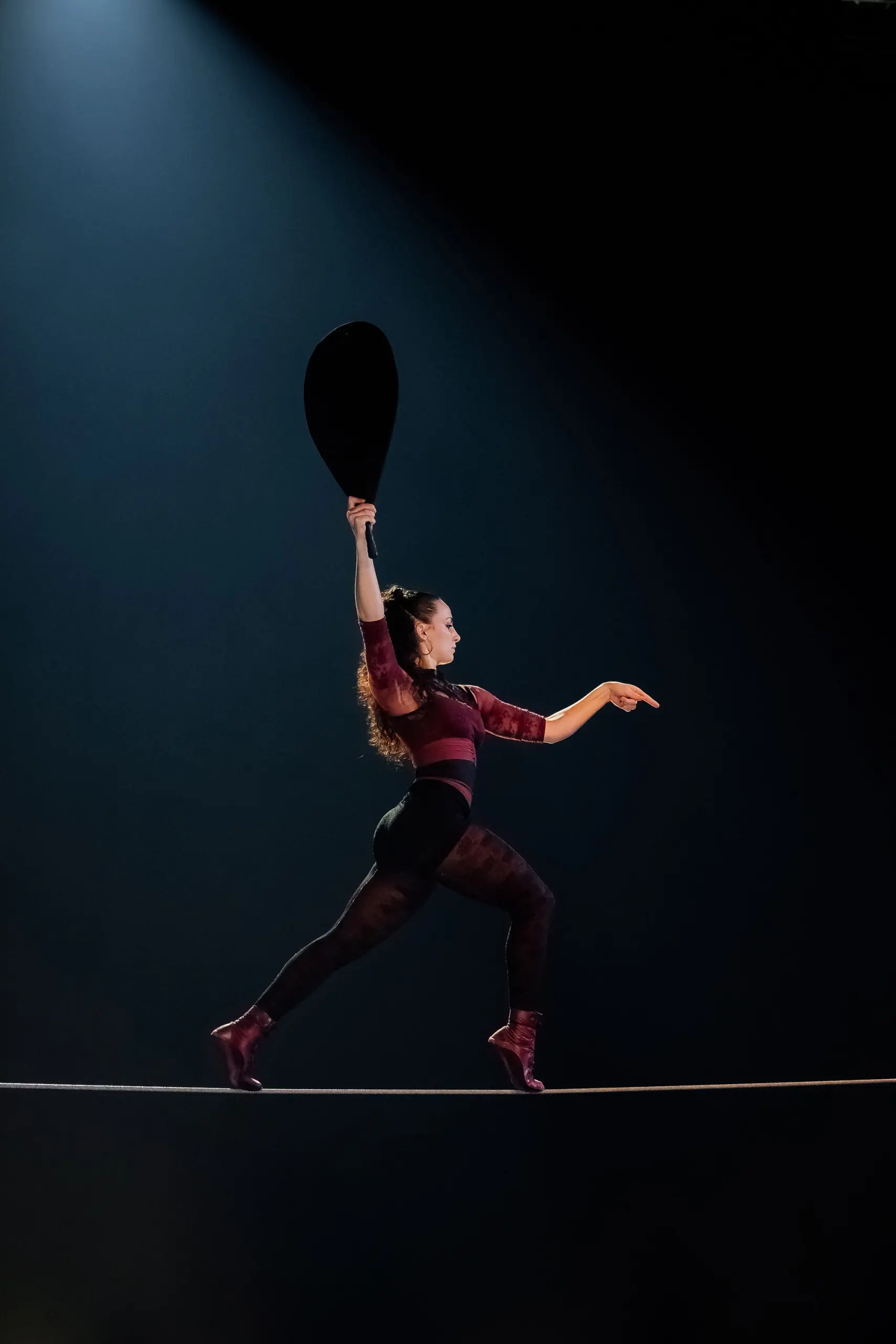 Rowan Allinson, vêtue de vêtements sombres et ajustés, se tient en équilibre sur une corde raide sous un projecteur, tenant un grand éventail noir d'une main et pointant vers l'avant de l'autre.