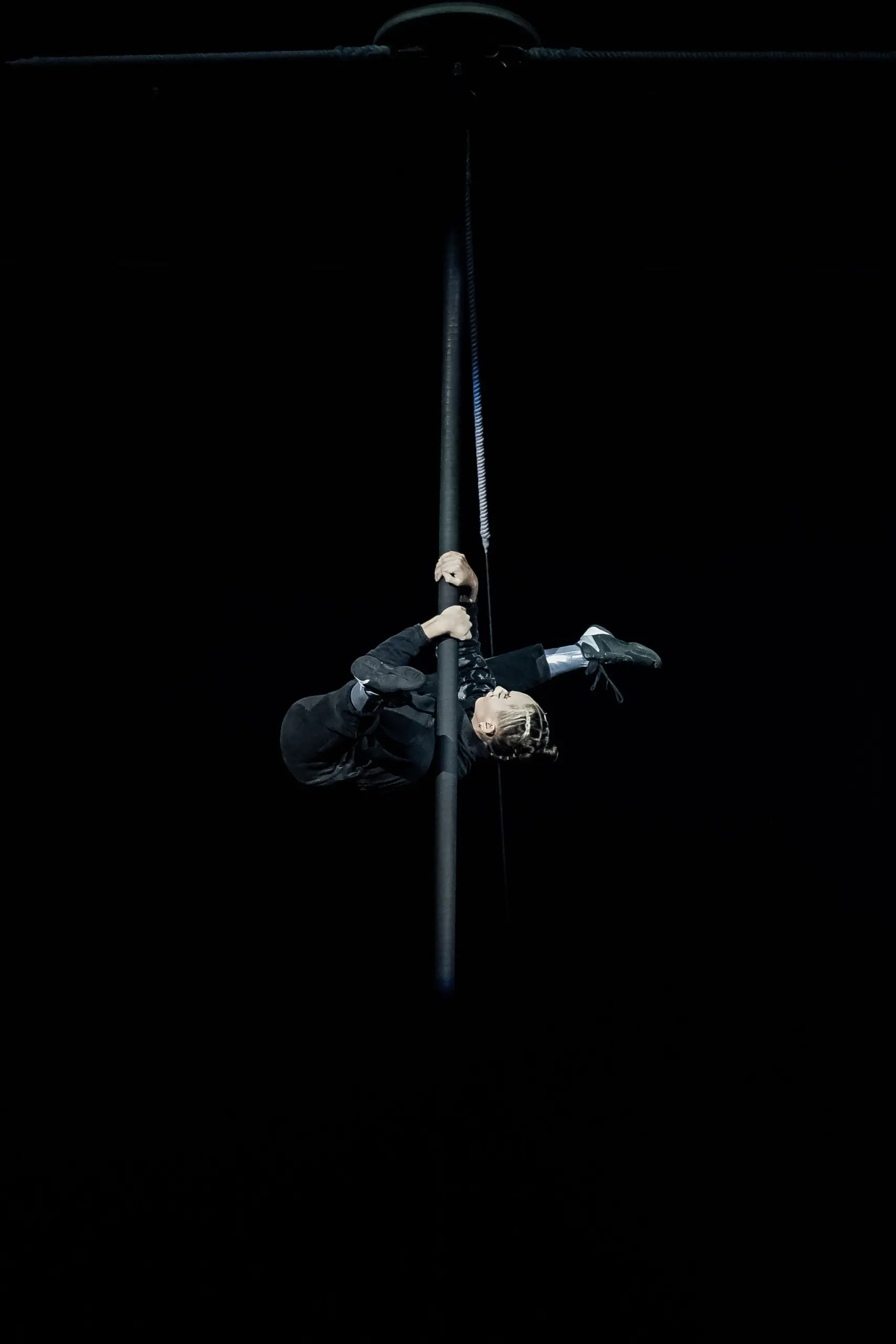Une personne vêtue d'un vêtement sombre exécute un mouvement acrobatique aérien en s'agrippant à un poteau vertical en hauteur - une pose saisissante connue sous le nom de béquille Maya - avec une jambe accrochée autour du poteau et la tête inclinée vers l'arrière, sur un fond sombre.