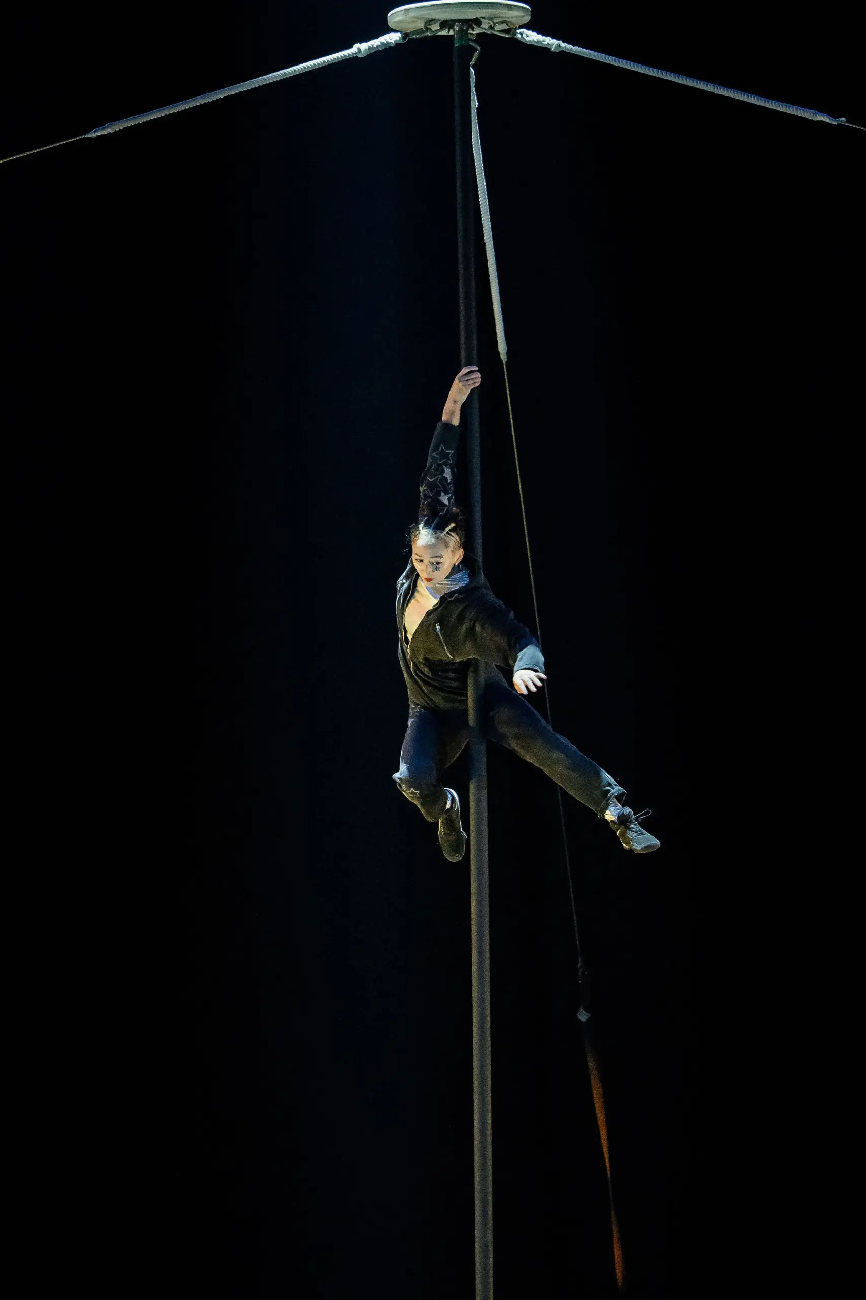 Un artiste de cirque vêtu de vêtements sombres est suspendu par un bras à un grand mât vertical, très haut au-dessus du sol, sur un fond sombre à l'éclairage dramatique - un acte qui rappelle les performances audacieuses de Maya Crutch.