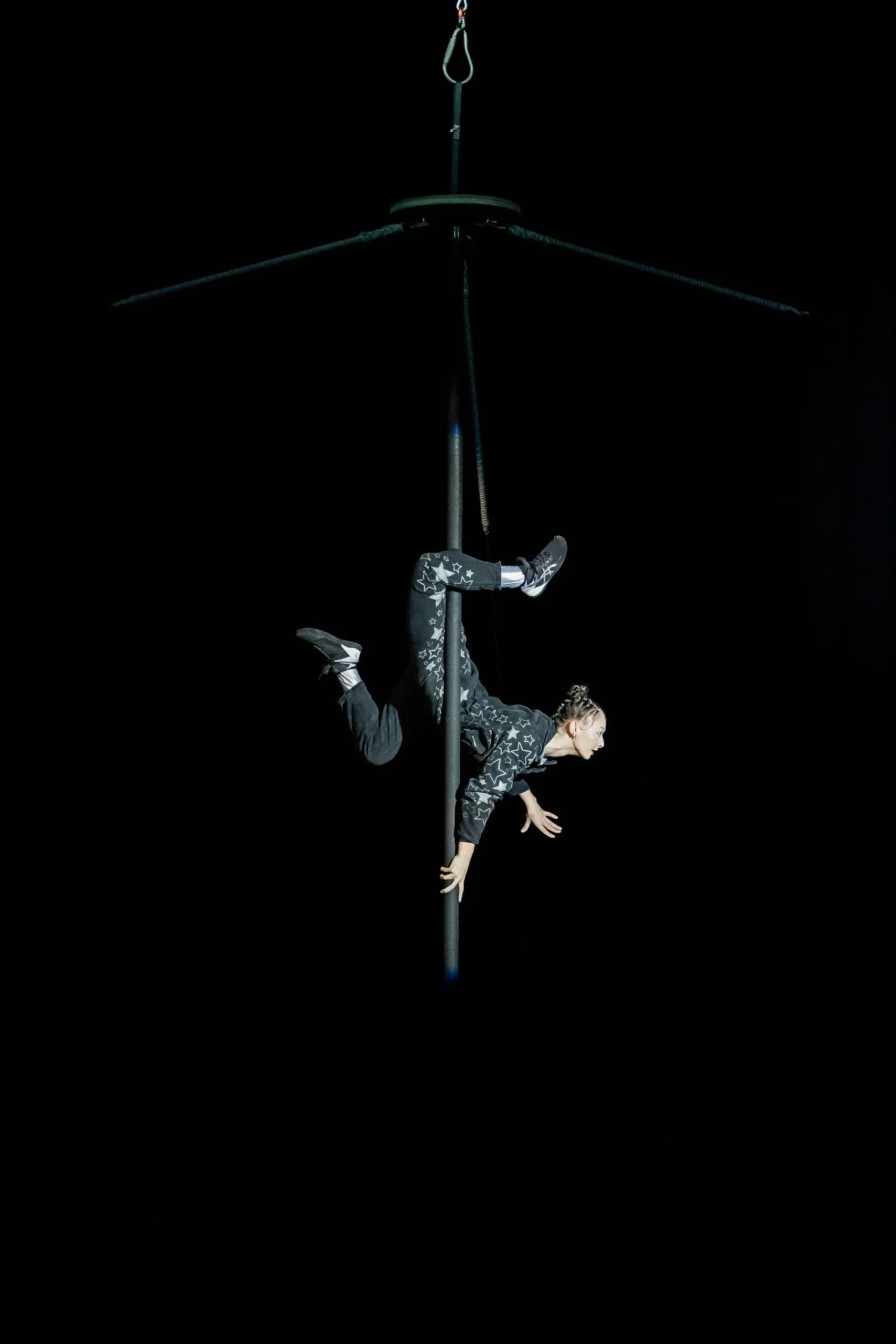 Une personne vêtue d'une tenue à motifs exécute une pose acrobatique aérienne, suspendue à l'envers à un poteau vertical dans l'obscurité, éclairée par un projecteur - capturant l'art audacieux de Maya Crutch.