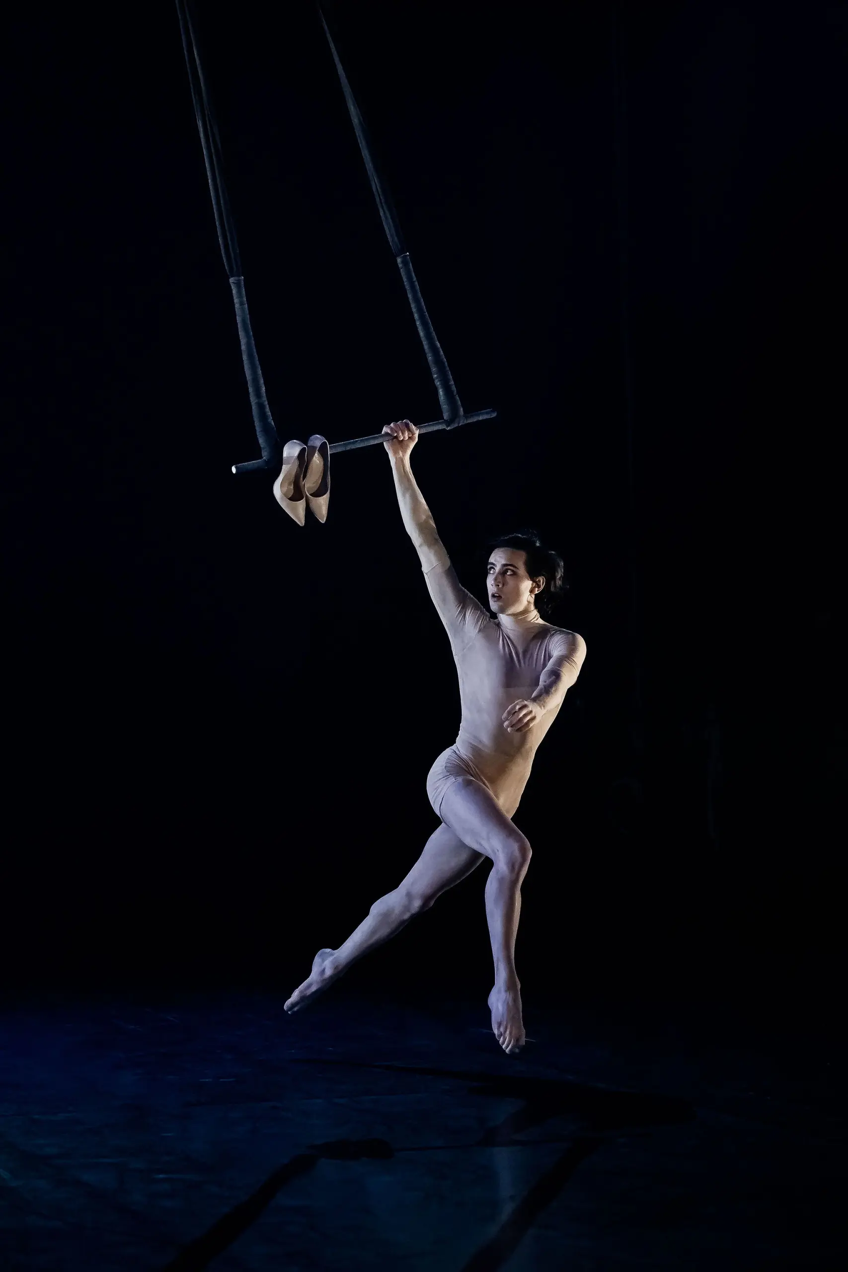 Une personne vêtue d'une tenue nude est gracieusement suspendue par un bras à un trapèze, ses chaussures de ballet reposant sur la barre, et se produit sur une scène sombre - un acte évoquant l'art de Manuel Gonzalo.