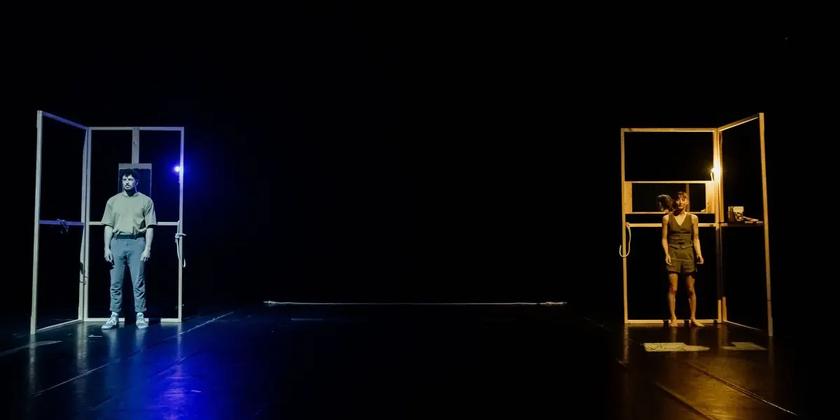 Deux personnes se tiennent à l'écart sur une scène sombre, chacune encadrée par une structure en bois distincte et éclairée par des lumières de couleurs différentes - bleu à gauche, jaune chaud à droite - soulignant leur isolement dans la mise en scène évocatrice de Clémence De Luca.