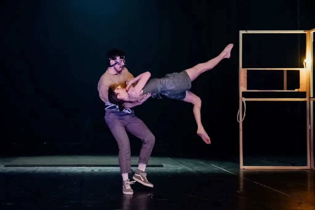 Un homme portant un masque soulève Clémence De Luca à l'horizontale dans une pose acrobatique sur une scène faiblement éclairée, à côté d'un cadre en bois avec une ampoule électrique.