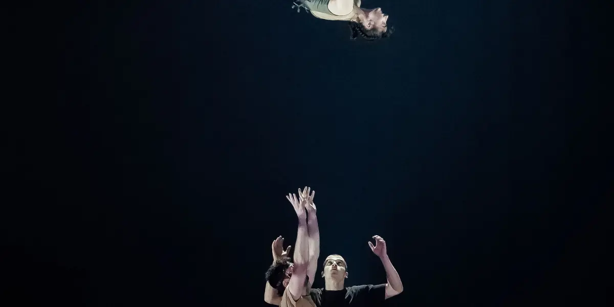 Un acrobate effectue un saut périlleux dans les airs au-dessus de deux artistes sur scène. Clémence De Luca est éclairée par un projecteur dramatique sur un fond sombre.