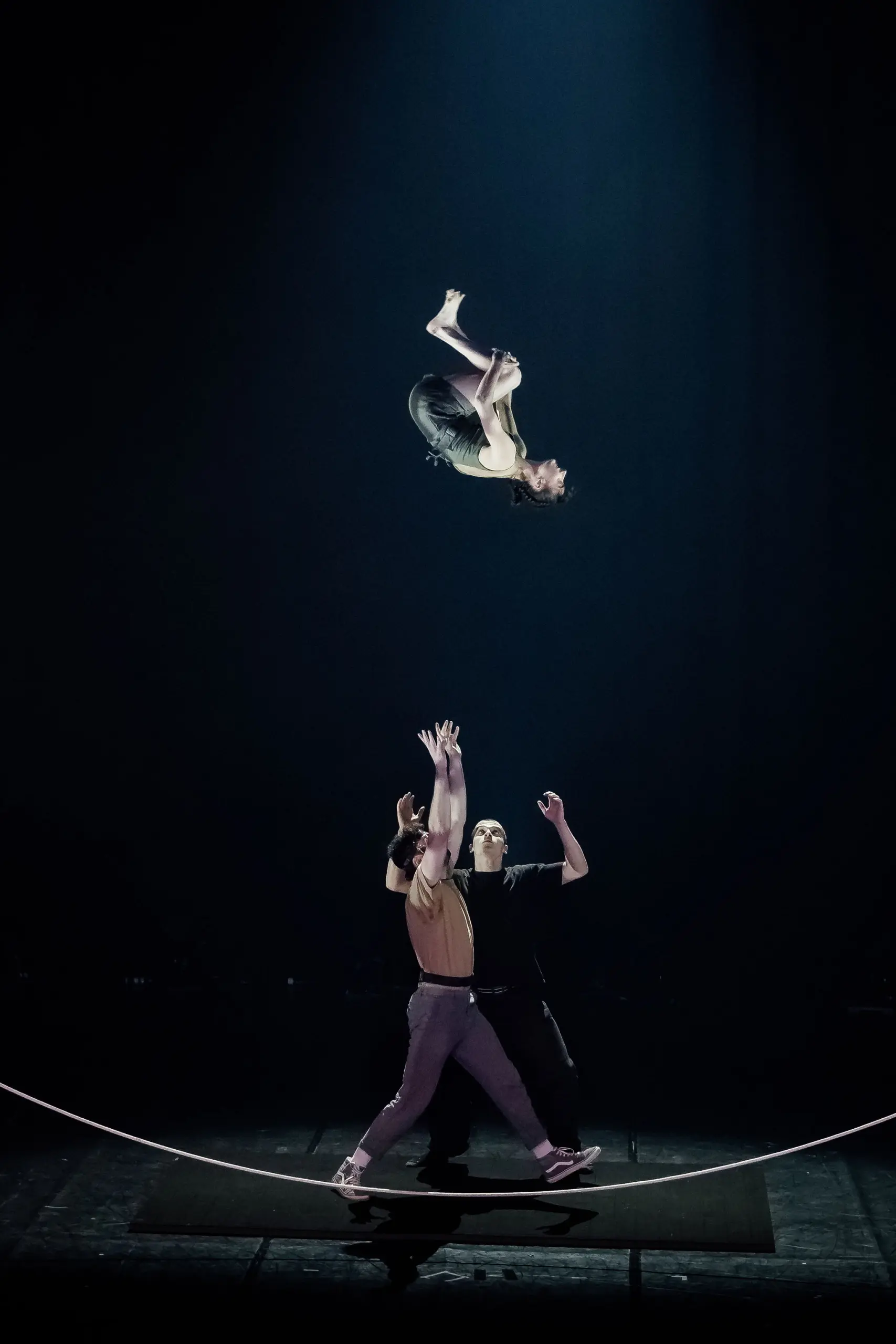 Un acrobate effectue un saut périlleux dans les airs au-dessus de deux artistes sur scène. Clémence De Luca est éclairée par un projecteur dramatique sur un fond sombre.