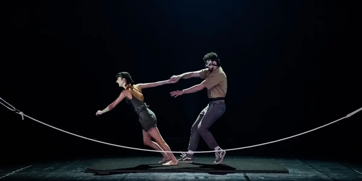 Deux artistes se tiennent en équilibre sur une slackline sur scène ; Clémence De Luca mène, penchée vers l'avant, tandis que l'homme la suit, s'agrippant à son bras pour la soutenir, tous deux éclairés par un projecteur sur un fond sombre.