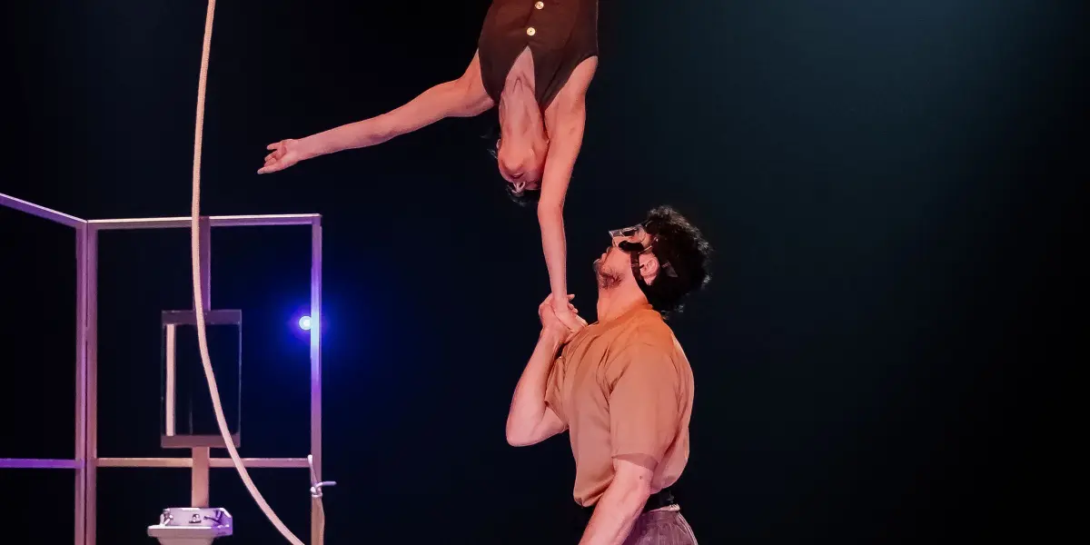 Deux artistes de cirque sur scène : l'un se tient en équilibre sur la main de Clémence De Luca tout en tenant une corde, les jambes tendues en fente. La scène est faiblement éclairée par des projecteurs et un minimum d'accessoires en arrière-plan.