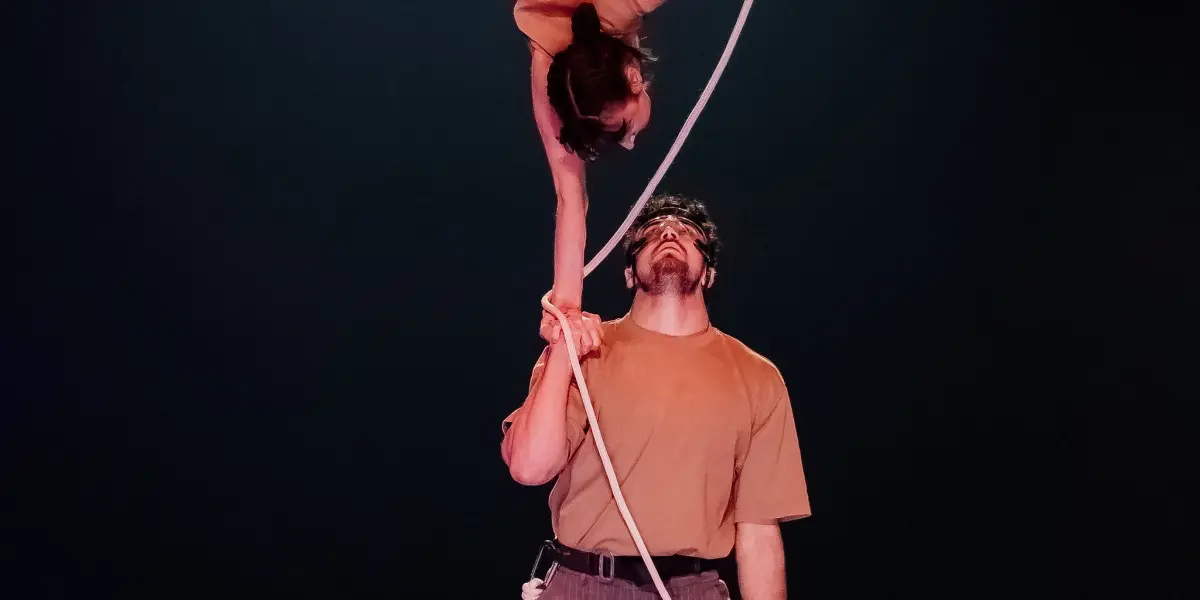 Un acrobate masculin se tient sur scène, faisant tenir Clémence De Luca en équilibre sur une main, la tête en bas, tandis qu'elle tient une corde blanche. La lumière tamisée met en valeur la force et l'équilibre des artistes.
