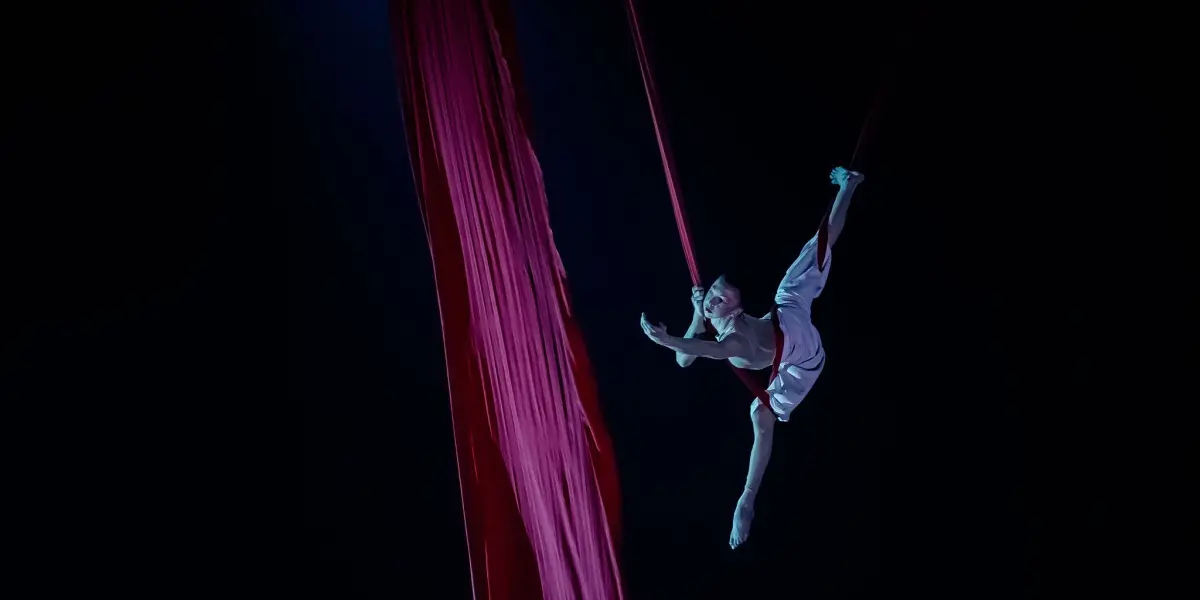 Arielle Gorodetskaya, dans un costume blanc, se suspend gracieusement à la soie aérienne rouge, étendant un bras et une jambe sur un fond sombre à l'éclairage dramatique.