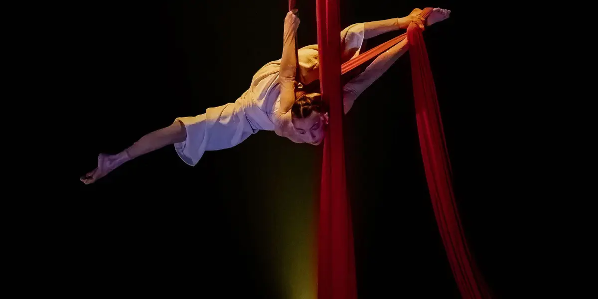 Arielle Gorodetskaya, une artiste aérienne, exécute une pose acrobatique en plein vol, suspendue à des soies rouges sur un fond sombre, éclairé par un projecteur.