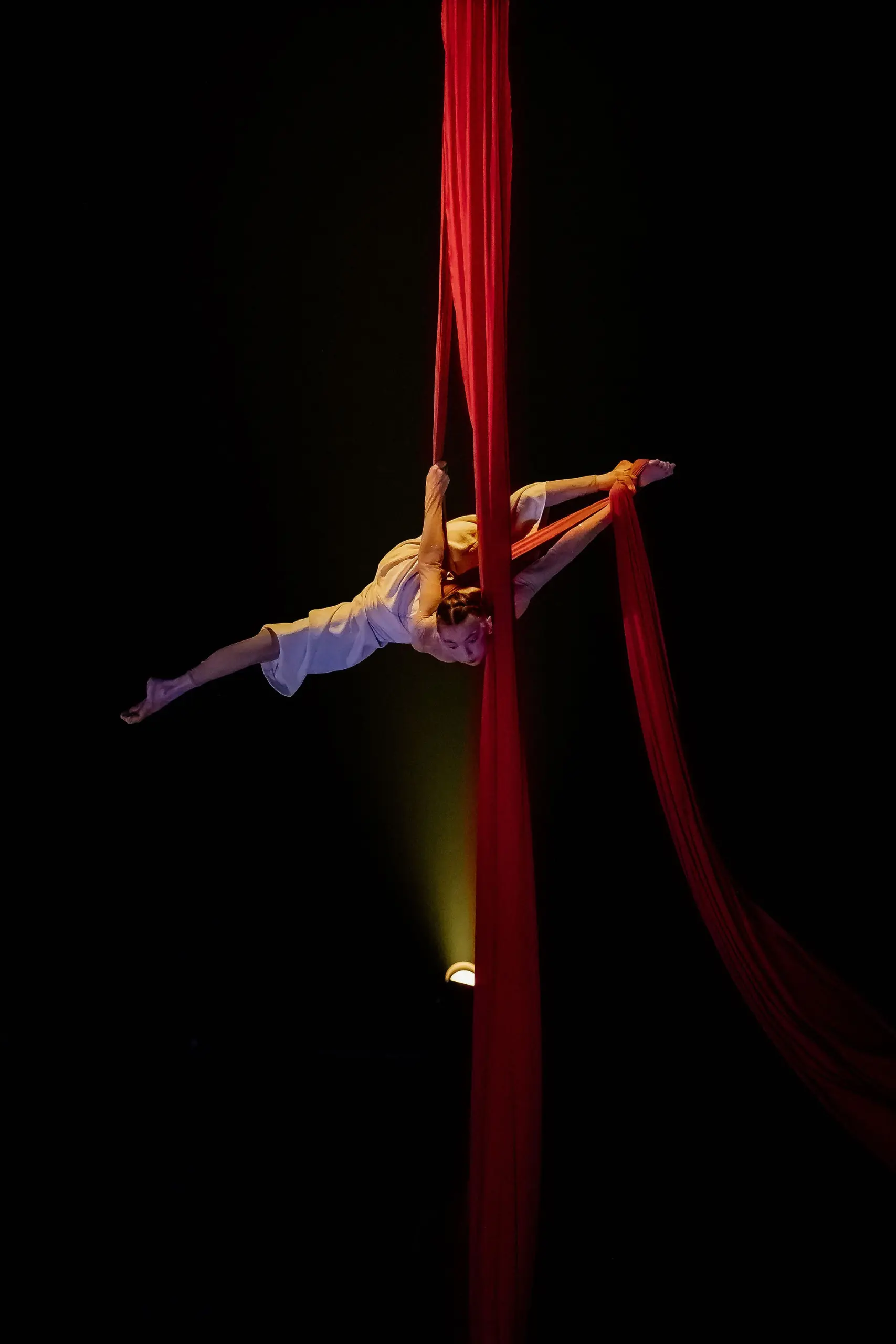 Arielle Gorodetskaya, une artiste aérienne, exécute une pose acrobatique en plein vol, suspendue à des soies rouges sur un fond sombre, éclairé par un projecteur.