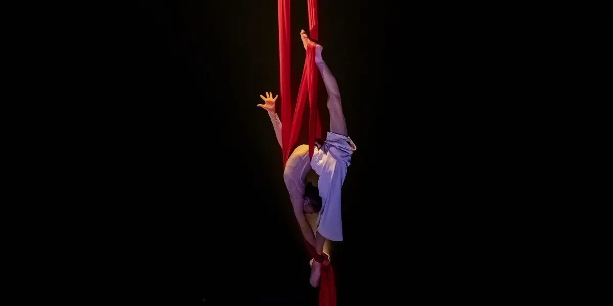 Arielle Gorodetskaya se produit en tant qu'artiste aérienne sur des soies rouges sur un fond sombre, avec une jambe tendue vers le haut et les bras et les jambes enveloppés dans le tissu, créant une pose dramatique et gracieuse.